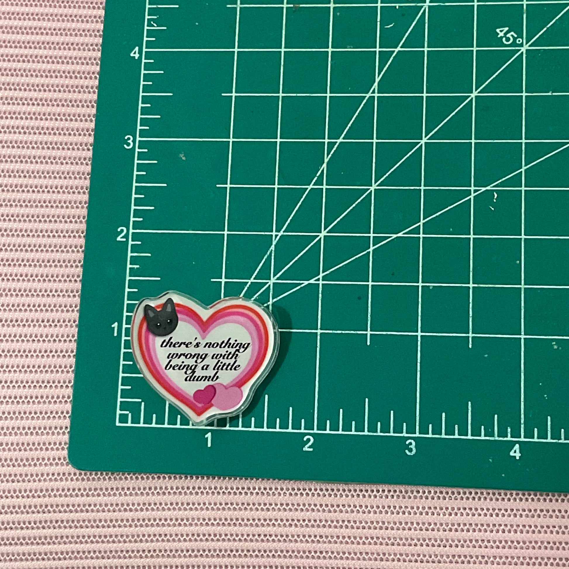 Heart-shaped pin with a cat design on a green cutting mat with grid lines.